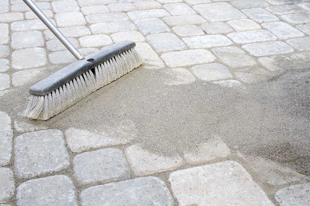 Person sweeping polymeric jointing sand into paver joints to prevent weed growth